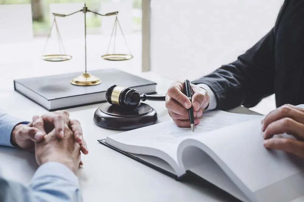 Lawyer signing documents with client, justice scales in background.