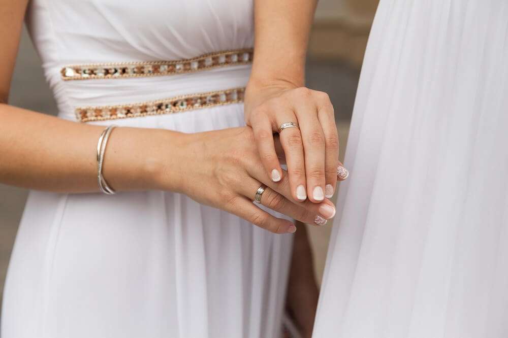 Two brides in white gowns displaying their wedding rings