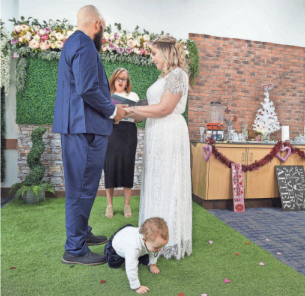 Couple exchanging vows while toddler plays on grass aisle.